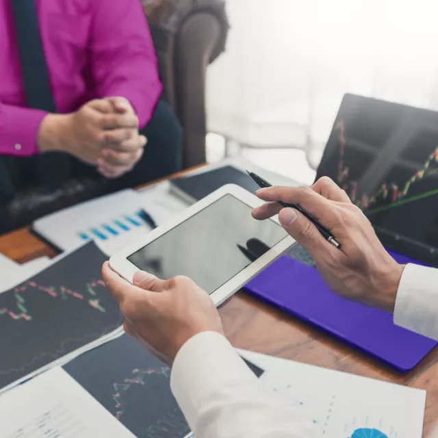 A man holding a tablet over a desk full of papers meeting with a woman about financials