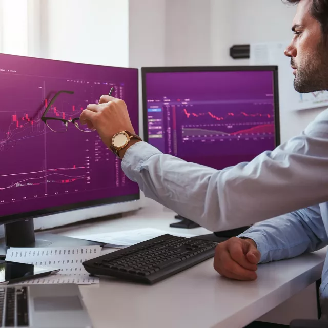 Image of man holding eyeglasses pointing at monitors with graphs
