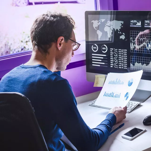 Image of man looking at paper with graphs, in front of computer showing graphs