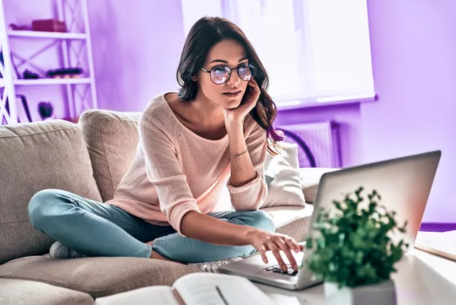 A person sitting cross-legged on a couch, using a laptop with a small potted plant nearby in a brightly lit room.