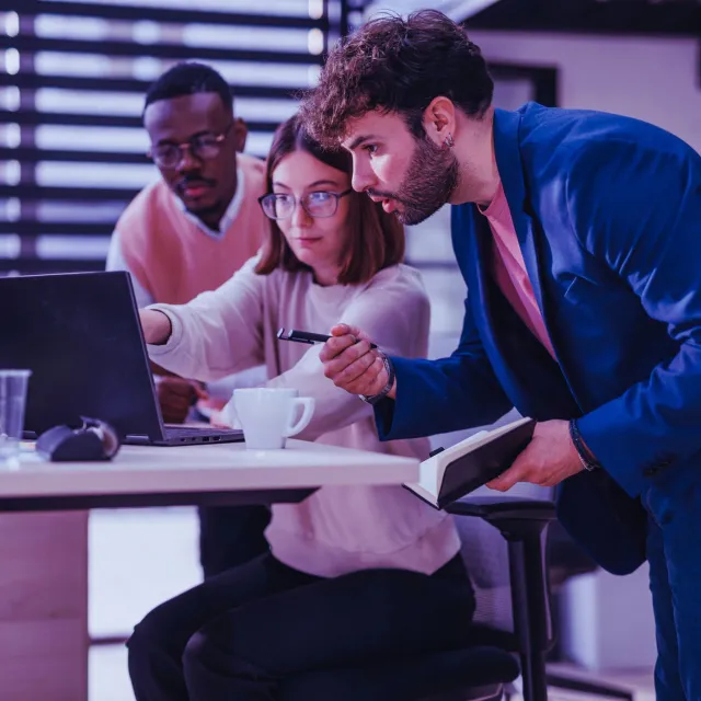 Three coworkers collaborating in a modern office, reviewing with a note and a laptop.