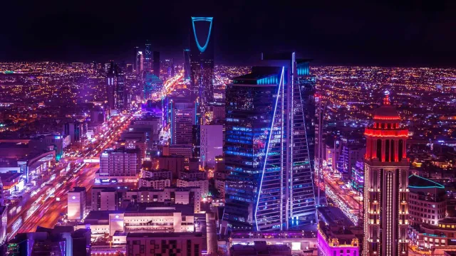 Night skyline of Riyadh with Kingdom Centre Tower lit in blue and bustling city lights.