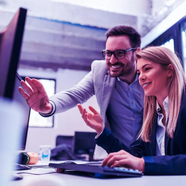 Two professionals in a modern office collaborating at a computer workstation, reviewing data on a large monitor.