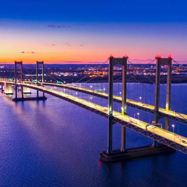 Suspension bridge illuminated at dusk spanning a wide river with city lights glowing in the background.