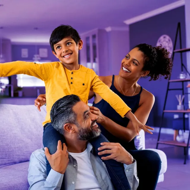 Family spending time together in a living room, with a child sitting on an adult’s shoulders while another person supports from behind.