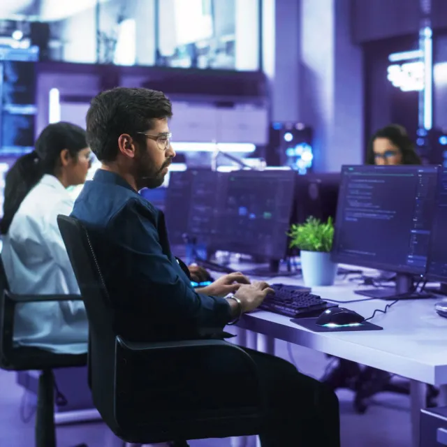 Cybersecurity operations center with IT professionals monitoring multiple screens displaying code and network data in a high-tech environment.