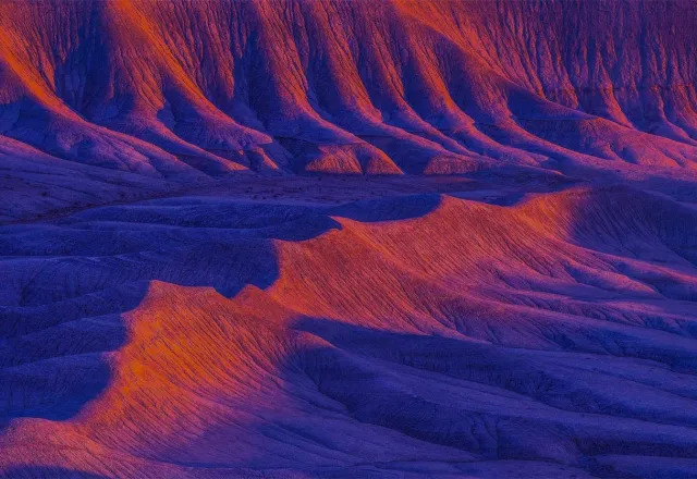 Colorful desert landscape with vibrant orange and purple ridges illuminated by dramatic sunset light.