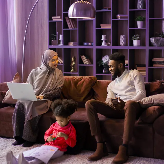 Family sitting on a couch in a modern living room with a laptop and bookshelves in the background.