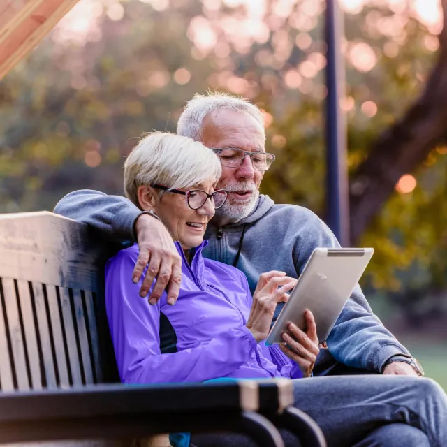 Older couple sitting on a park bench using a digital tablet.