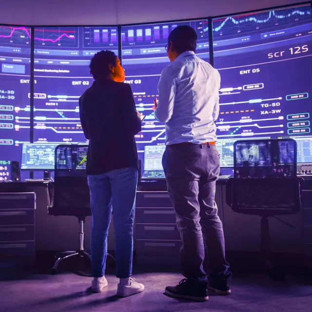 In the System Control Room Project Manager and IT Technical Engineer Have Discussion, they're surrounded by Multiple Monitors with Graphics. Big Monitor Shows Interactive Server Blockchain Info.
