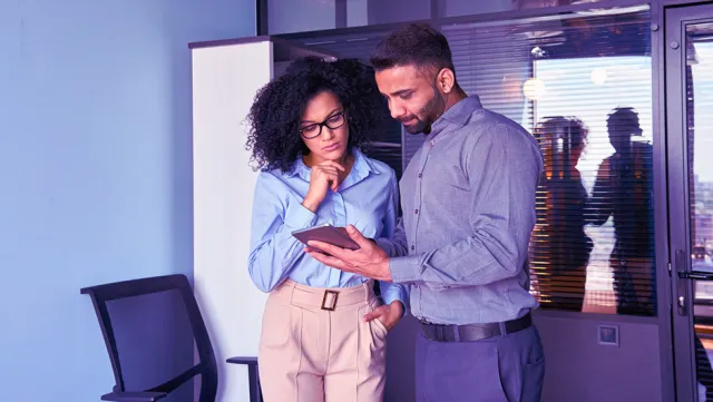 Two professionals standing in an office reviewing information on a digital tablet.