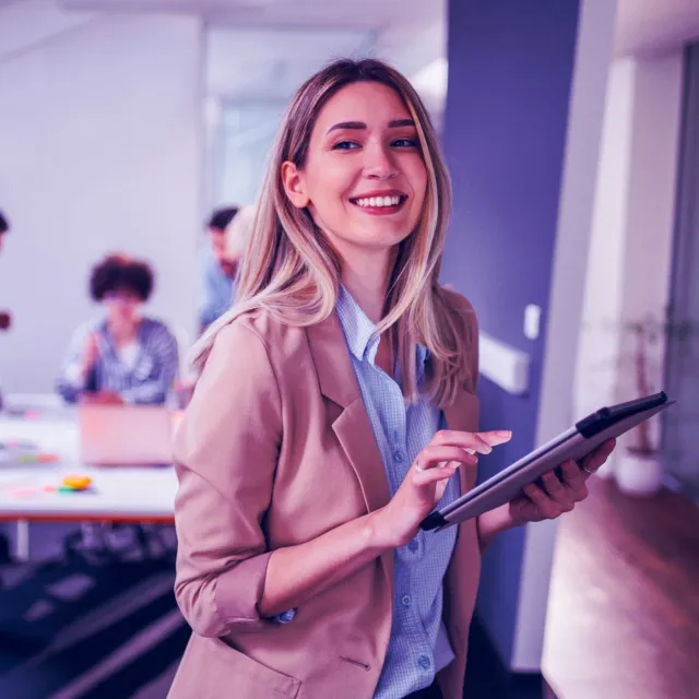 Professional using a digital tablet in a modern office with team collaborating in the background.