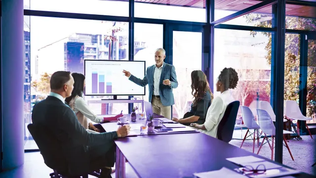 Business team meeting in a modern conference room with a presenter explaining data charts on a digital screen during a strategy discussion.
