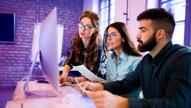 A group of three people collaborating at a desk, reviewing documents and working together on a large desktop computer in a modern office setting.