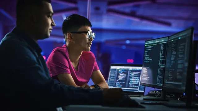 Two people working together at a desk, reviewing code on multiple computer monitors in a dimly lit workspace.