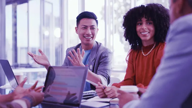 A group of colleagues engaged in a discussion around a table with laptops and coffee cups.