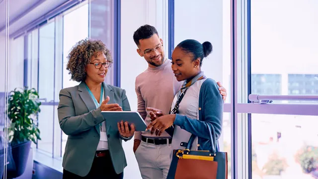 A small group of colleagues standing in a hallway, discussing information on a tablet.