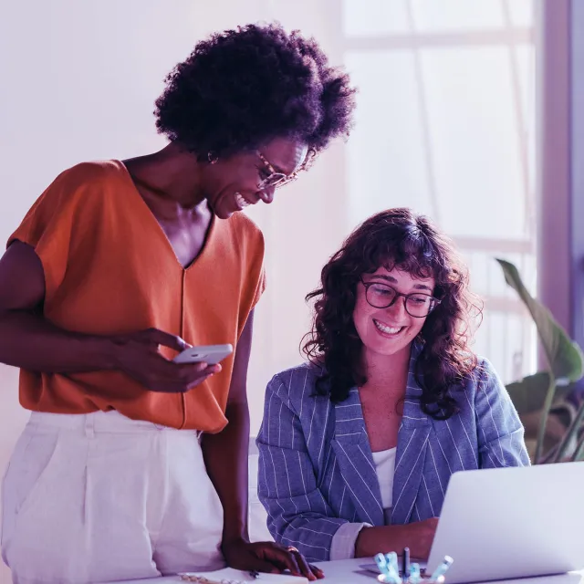 Two colleagues collaborate in a bright office; one stands holding a smartphone while the other sits at a desk working on a laptop.