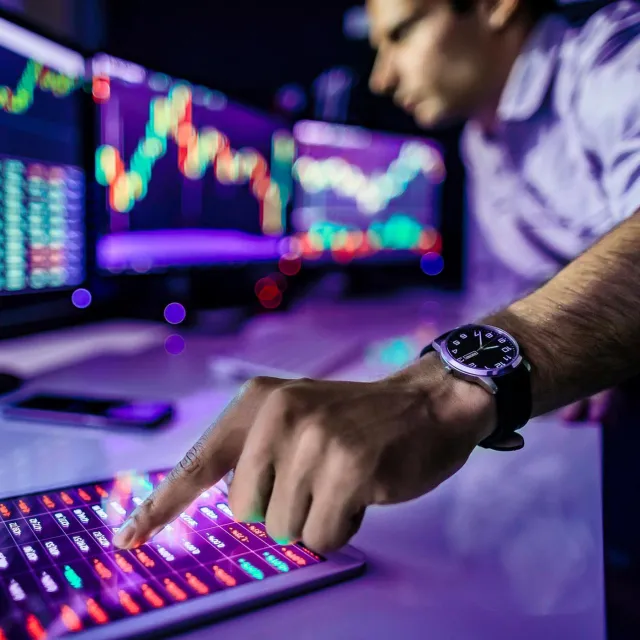 Hand tapping a tablet displaying live market data with stock charts on monitors in the background.