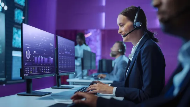 Professionals in a high-tech control room with purple lighting, working at computer monitors displaying data analytics and graphs, wearing headsets for communication and monitoring.