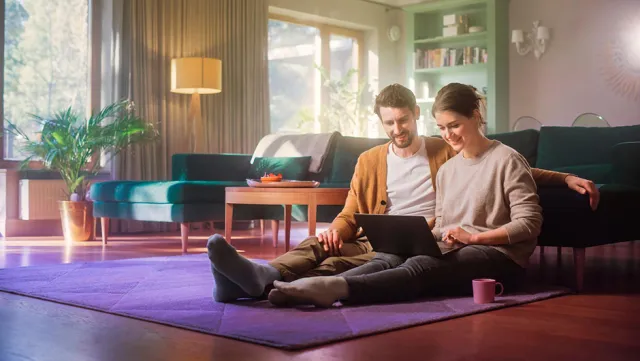 A couple sits on the living room floor using a laptop in a bright, cozy space.