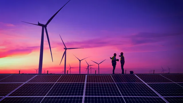 Two people on a solar panel field with wind turbines in the background.
