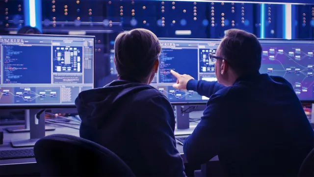 Two Professional IT Programers Discussing Blockchain Data Network Architecture Design and Development Shown on Desktop Computer Display. Working Data Center Technical Department with Server Racks