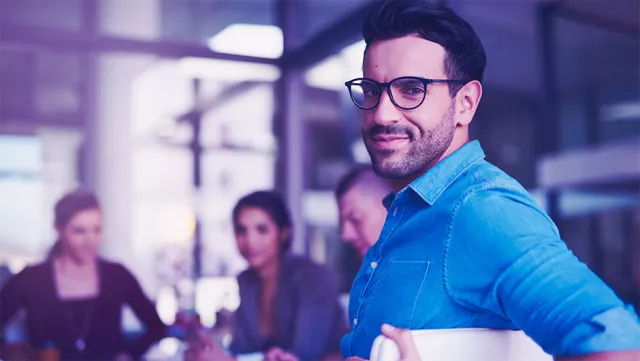 Confident professional holding laptop in a modern office with collaborative team in background