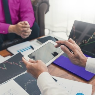 A man holding a tablet over a desk full of papers meeting with a woman about financials