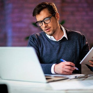 Image of a male looking at a laptop while holding a pen and a tablet