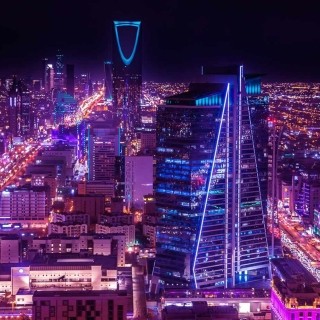 Night skyline of Riyadh with Kingdom Centre Tower lit in blue and bustling city lights.