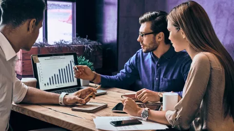 Three people at an office table, looking at a laptop with a graph