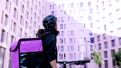 A delivery person with a large purple delivery bag stands next to a bicycle, looking up at a tall apartment building in an urban setting.