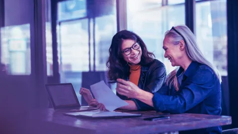 Two people collaborating over documents at a table with a laptop in a modern office setting.